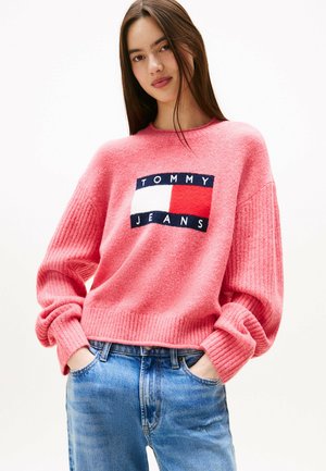 Giovane donna con lunghi capelli castani che indossa un maglione rosa di Tommy Jeans e jeans blu, mani in tasca, su uno sfondo chiaro e semplice.