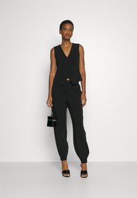 Gilet noir sans manches avec un décolleté en V et un léger drapé, associé à un pantalon noir fuselé avec des poignets élastiques et des poches latérales.