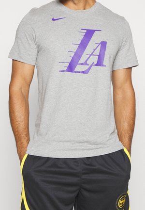 Homme portant un t-shirt Nike gris avec un grand logo "LA" violet et un short noir avec bordure et emblème jaunes, debout les mains dans les poches.