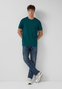 Teal ronde hals t-shirt van katoenmix, gecombineerd met blauwe jeans en lichte sneakers. Model staat ontspannen met de armen langs het lichaam tegen een effen achtergrond.