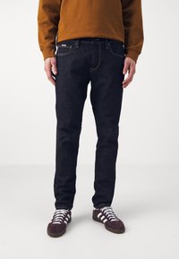 Tmavě modré džíny s úzkým střihem, kontrastním žlutým prošitím a pěti kapsami. Nosené s hnědou mikinou a bordó teniskami.