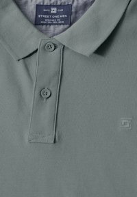 Camisa polo de tela verde claro con un acabado texturizado, que presenta un cuello clásico, dos botones y un parche con el logo cerca del dobladillo.