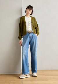 Olivgrüner Wollcardigan über einem weißen Shirt mit Knöpfen, kombiniert mit hochgeschnittenen blauen Jeans und beigen Sneakers, steht vor einer neutralen Wand.