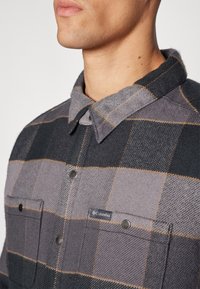 Grå rutete skjorte med knappelukking, som har to frontlommer og krage. Stoffet har en teksturert overflate og subtile oransje detaljer.