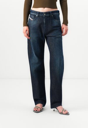 Femme portant un jean droit bleu foncé taille haute, un pull côtelé marron court et des sandales à talons argentées à brides.