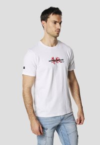 T-shirt bianca in cotone con maniche corte e scollo rotondo, con una scritta nera e rossa sulla parte anteriore e un piccolo logo sulla manica.