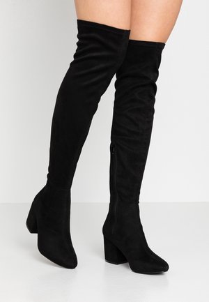 Des jambes portant des bottes en suede noir au-dessus du genou avec des talons carrés et des zips sur les côtés, se tenant sur un sol blanc contre un fond clair.