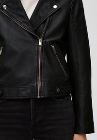 Schwarze Leder-Motorradjacke mit silberner Hardware, asymmetrischen Reißverschlüssen und Druckknopf-Akzenten. Glatte Textur, verkürzte Passform und taillierter Schnitt.