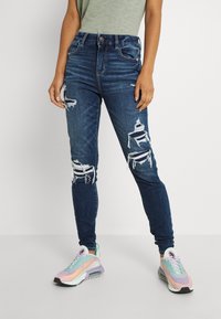 Högmidjade, mörkblå jeans i denim med slitna detaljer vid knäna och en slim fit, tillsammans med flerfärgade sportskor.