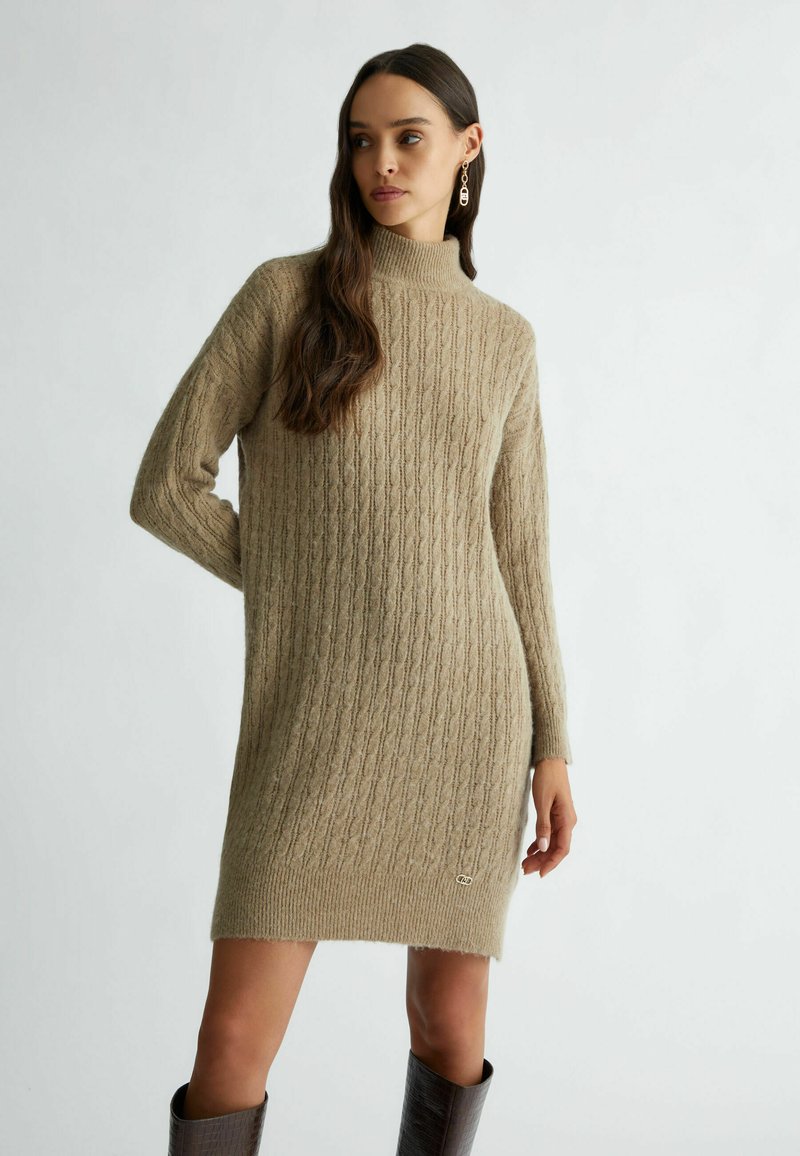 Abito in maglia marrone chiaro, con collo alto, texture a coste e dettaglio del logo sull'orlo, dotato di maniche lunghe.
