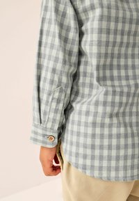 Enfant portant une chemise à manches longues à carreaux gris clair et blancs avec un pantalon beige, vue du poignet et de l'ourlet latéral.