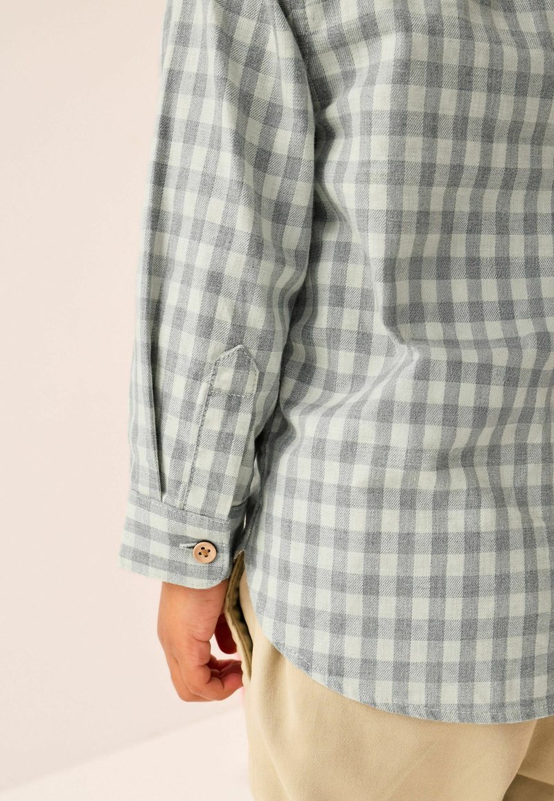 Enfant portant une chemise à manches longues à carreaux gris clair et blancs avec un pantalon beige, vue du poignet et de l'ourlet latéral.
