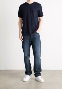 Camiseta de algodón azul marino con mangas cortas, cuello redondo y pequeño logo en el pecho, combinada con jeans de mezclilla azules y zapatillas blancas.
