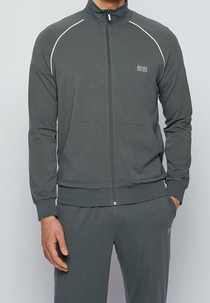 Man draagt een donkergrijs trainingsjack met rits en bijpassende broek, met het logo "BOSS HUGO BOSS" op de linkerborst en de linker bovenbeen.