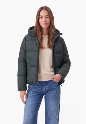 Femme portant une doudoune à capuche vert foncé sur un pull beige et un jean bleu, debout avec une main dans la poche sur fond blanc.