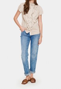 Beige Rüschenbluse mit kurzen Ärmeln, kombiniert mit hellblauen Jeans mit umgeschlagenem Saum und braunen Riemensandalen.