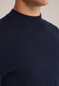 Man som bär en mörk marinblå ribbad tröja med hög hals, kort i modellen för att visa halsen och övre bröstet mot en enkel bakgrund.