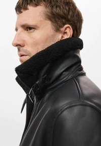 Chaqueta de cuero negra con un grueso cuello de borrego negro, que presenta un cierre de cremallera y costuras detalladas a lo largo de los hombros y el cuello.