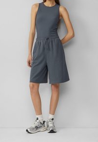 Grijze geribbelde tanktop gecombineerd met grijze shorts met een elastische tailleband. Draagt zilveren sportschoenen en witte sokken. Simpel ontwerp.