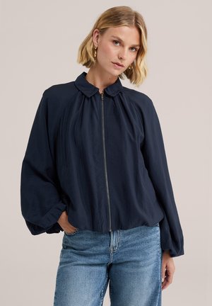 Femme aux cheveux blonds courts, portant des boucles d'oreilles créoles dorées, une veste zippée bleu marine foncé avec des manches ballon et un jean bleu clair.