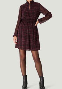 Langärmliges Kleid mit hohem Kragen, strukturiertem Stoff im dunkelroten und schwarzen Karomuster, taillierter Taille und ausgestelltem Rock. Kombiniert mit Stiefeletten.