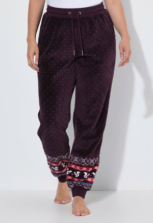 Mujer con pantalones de pijama de color púrpura oscuro con pequeños puntos y puños estampados y coloridos con ardillas, de pie descalza sobre un fondo blanco.