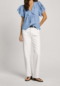 Blusa de algodón azul claro con mangas con volantes, patrón decorativo de ojales, escote en forma de V y botones. Combinada con jeans rectos blancos y sandalias negras.