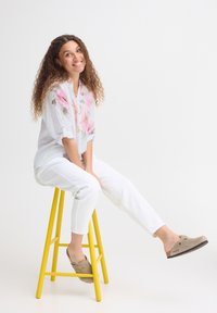 Jeune femme aux cheveux bouclés souriante, assise sur un tabouret jaune, portant un pantalon blanc, une blouse fleurie et des chaussures beige sans lacets.