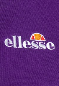 Tissu violet avec logo « ellesse » brodé en blanc et un motif en demi-cercle orange et rouge au-dessus du texte.