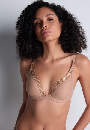 Femme aux cheveux bouclés portant un soutien-gorge couleur chair avec une bordure en dentelle, posant devant un fond gris clair uni.