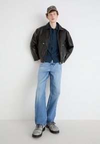Veste en cuir noir avec poches zippées, chemise noire à boutons, jean évasé bleu clair et chaussures grises avec lacets noirs.