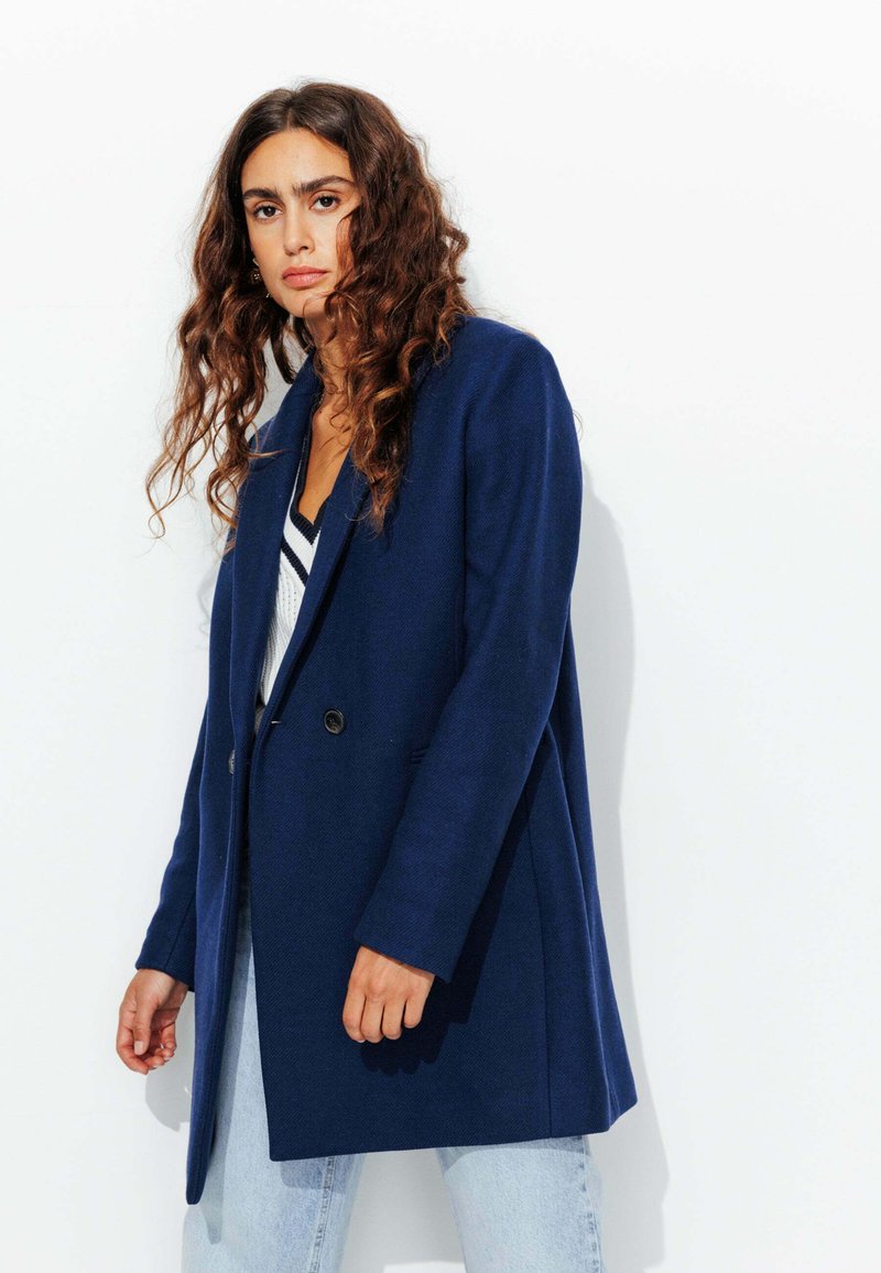 Blazer bleu marine avec un col cranté, fermeture à un bouton et deux poches latérales. Porté par-dessus un pull rayé blanc et bleu marine.