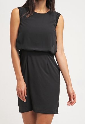 Femme portant une robe noire sans manches, longueur genou, avec un haut ample et une jupe ajustée, sur un fond clair uni.