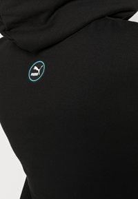 Sudadera negra con una textura suave, que presenta una capucha redondeada y un logotipo circular de Puma en turquesa y blanco en la parte posterior.