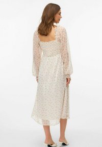 Robe longue et transparente en crème avec un délicat imprimé floral rose, corsage smocké et manches bouffantes. Elle présente un dos carré et un ourlet mi-long.