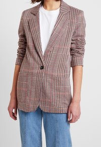 Blazer i röd, svart och vit rutmönster; har en enda knapp, långa ärmar och två framfickor. Bärs över en vit t-shirt.