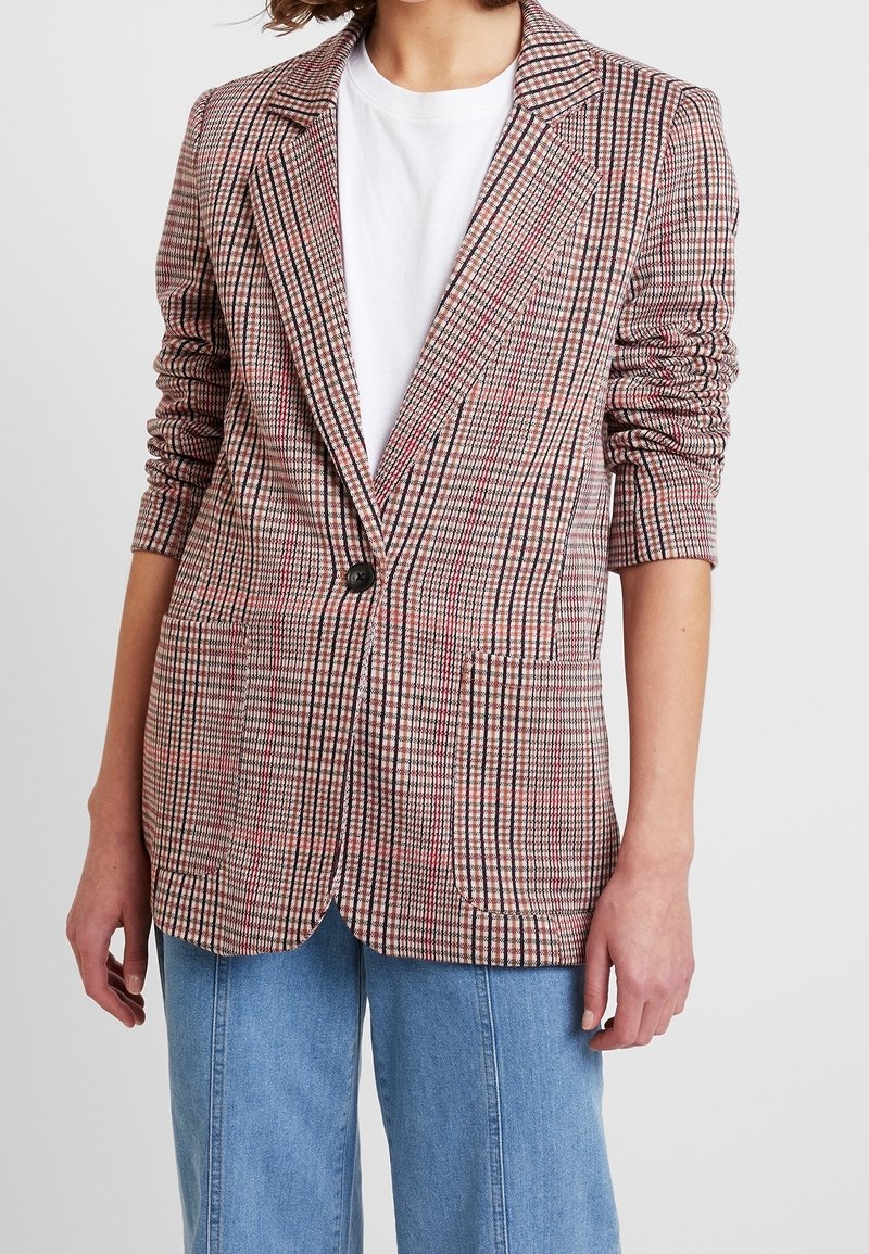 Blazer i röd, svart och vit rutmönster; har en enda knapp, långa ärmar och två framfickor. Bärs över en vit t-shirt.