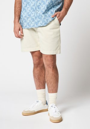 Mann trägt cremefarbene, strukturierte Shorts, weiße Socken, weiße Turnschuhe mit beigefarbenen Sohlen und ein blaues Hemd mit Blumenmuster, eine Hand in der Hosentasche.