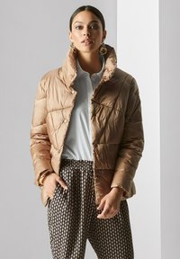 Beige Steppjacke mit hohem Kragen, gesteppter Optik und Druckknöpfen, kombiniert mit einer hellen Bluse und gemusterten Hosen.