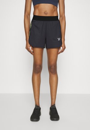 Mujer con pantalones cortos deportivos negros con una cintura ancha, zapatillas negras y un top deportivo corto oscuro, de pie frente a un fondo liso.