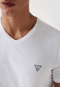 T-shirt bianco con scollatura a V realizzato in tessuto liscio, caratterizzato da un piccolo logo triangolare nero nella parte sinistra del petto.