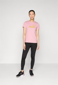 Rožinė medvilninė marškinė su auksiniu "PUMA" logotipu, derinama su juodais leggingais ir juodais sportiniais batais. Paprastas dizainas, trumpomis rankovėmis ir prigludusia forma.