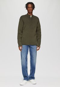 Khaki-grüner gerippter Pullover mit hohem Kragen und Reißverschluss, kombiniert mit hellblauen geraden Jeans und weißen Sneakers. Einfaches, strukturiertes Design.