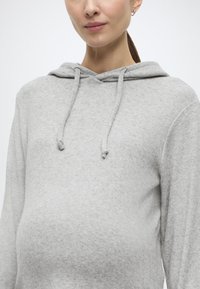 Gaiši pelēks džemperis ar kapuci, izgatavots no mīksta auduma, ar kanguropodi, auklu kapucē un brīvu piegriezumu, kas paredzēts komfortam grūtniecības laikā.