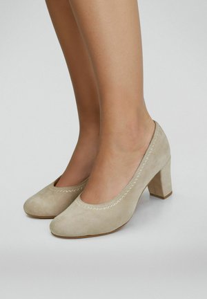 Chaussures à talons blocs à bout rond en daim beige portées pieds nus, vues de profil sur un fond neutre.