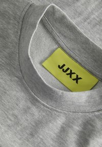 Camiseta de algodón gris con cuello redondo y un collar interno texturizado. Presenta una etiqueta amarilla brillante con "JUXX" impreso en negro.