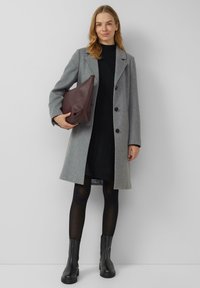 Grauer Wollmantel mit schwarzen Knöpfen, getragen über einem schwarzen Kleid. Dunkle Strumpfhosen und schwarze Stiefeletten, dazu eine bordeauxrote Lederhandtasche.