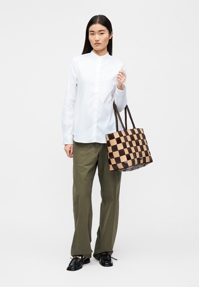 Καφέ και μπεζ τσάντα ώμου με καρό σχέδιο και υφαντή υφή, δύο χερούλια και επίπεδη βάση. Μοντέλο φορεί λευκό πουκάμισο με κουμπιά και χακί παντελόνι.