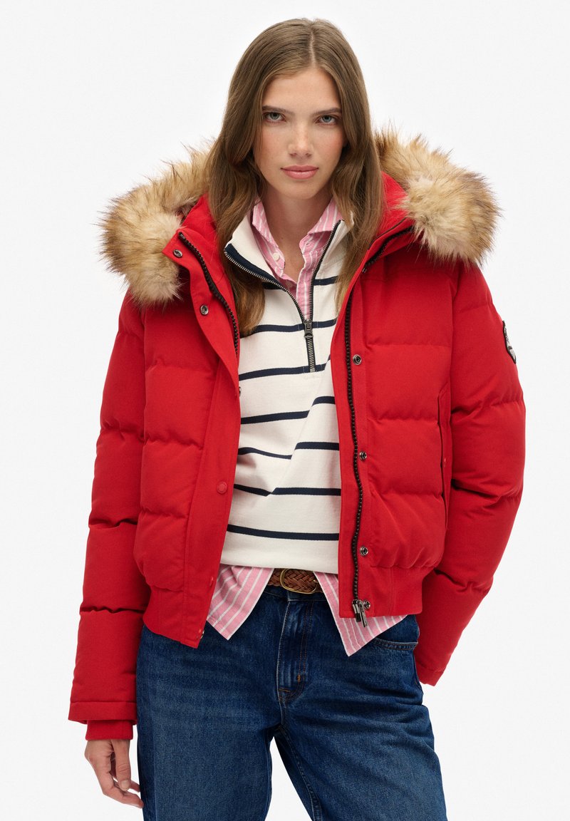 Junge Frau trägt eine rote Steppjacke mit Fellkapuze, gestreiften weißen und marineblauen Pullover, rosa Hemd und blaue Jeans vor weißem Hintergrund.