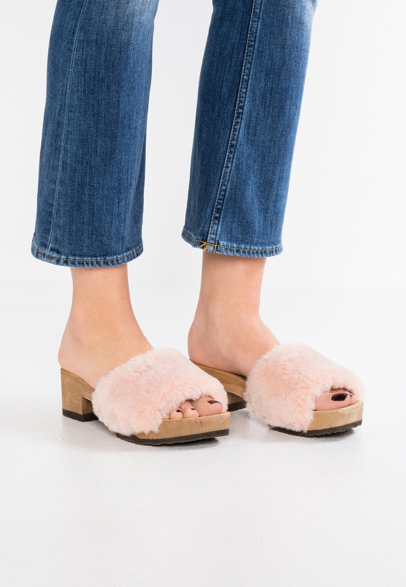 Růžové chlupaté pantofle s dřevěným blokovým podpatkem, s otevřenou špičkou a jemnou texturou. Nosené s deninovými kalhotami.